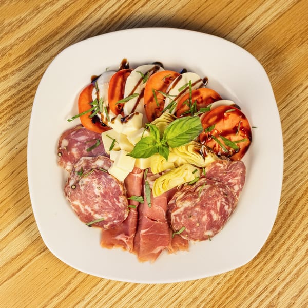 White plate with caprese salad, salami, prosciutto, cheese cubes, and artichoke hearts on a wooden table.
