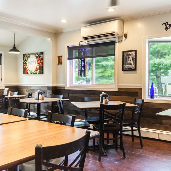 Empty restaurant dining room featuring wooden tables, dark chairs, wood paneling, and bright windows.
