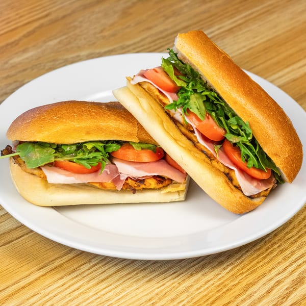Two gourmet sandwiches with prosciutto, tomatoes, and arugula on a white plate.
